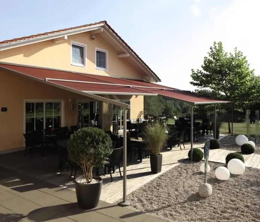 Haus mit roter Pergola-Markise über Terrasse mit Sitzplätzen und Pflanzen.