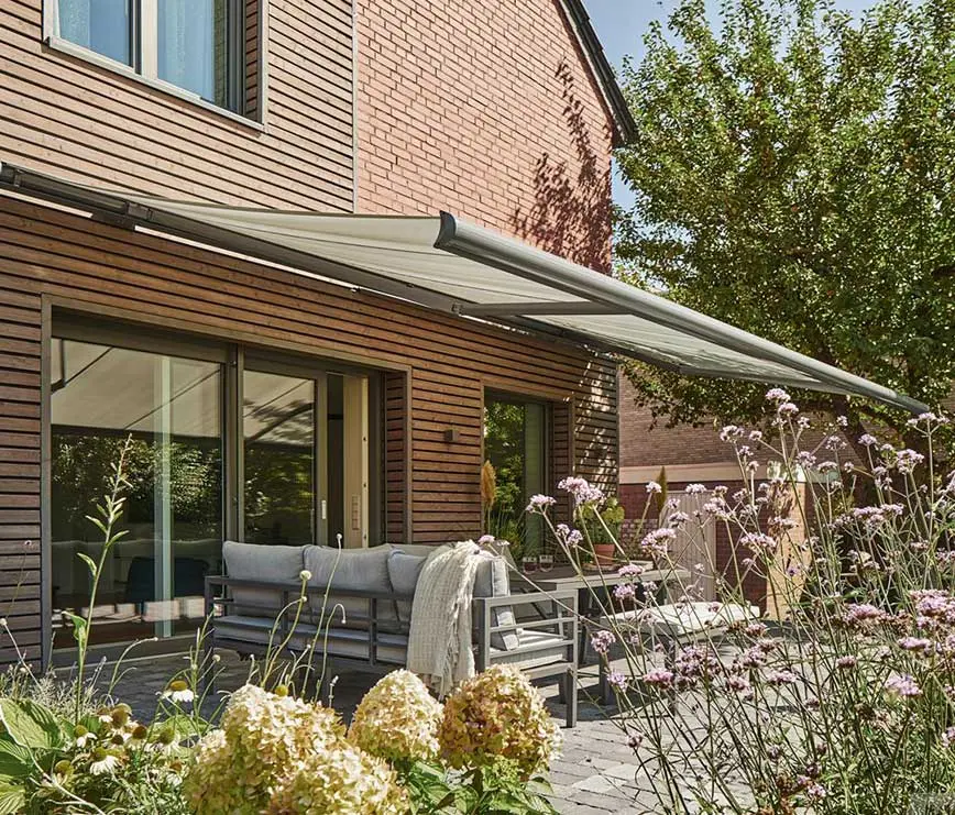 Moderne Terrasse mit Markise, Holzfassade und blühendem Garten im Sonnenlicht.